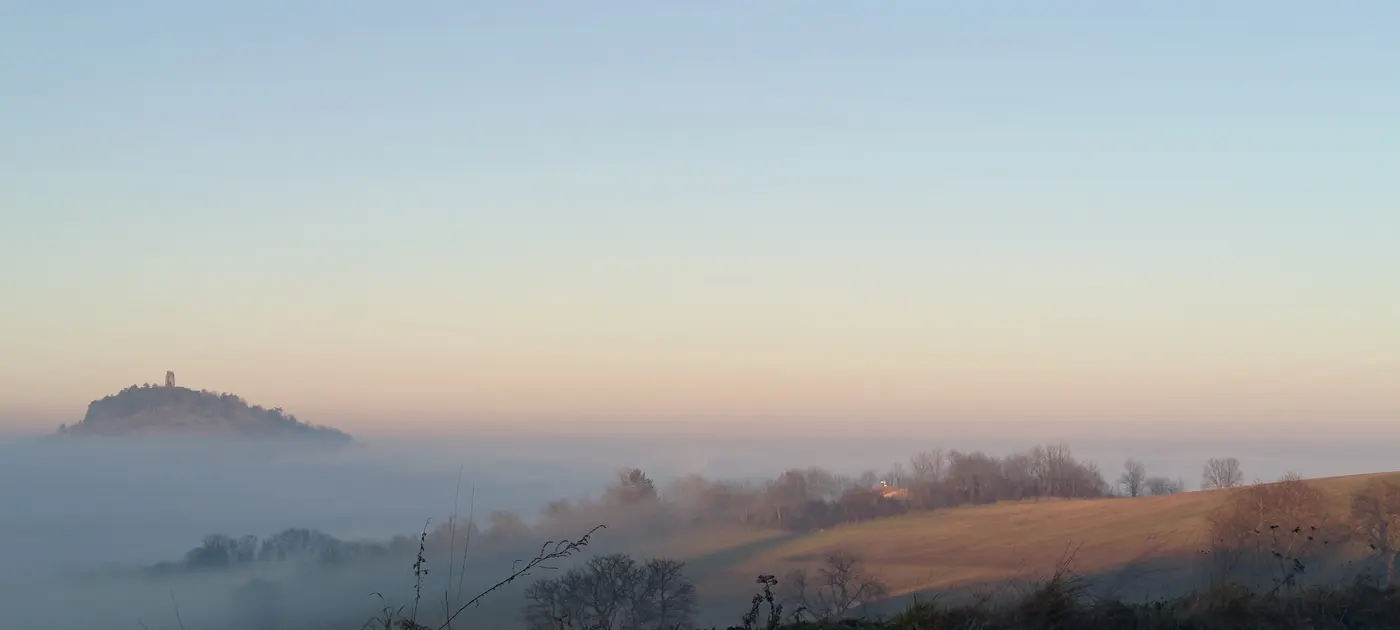 La butte à la tour, à gauche, posée sur la mer de brume ; à droite un versant de prés déjà touché par le soleil rasant, avec ses haies givrées et ses lignes électriques.