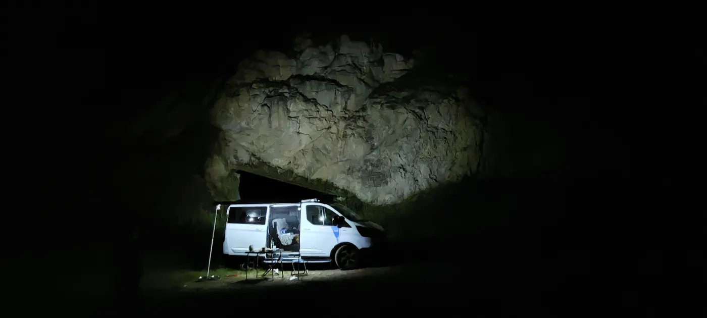 En pleine nuit, un fourgon aménagé garé sous un surplomb rocheux, éclairé par deux lampes sur pied qui révèlent la voûte de calcaire au-dessus de lui ; une table et deux sièges pliants sont installés devant la porte latérale.