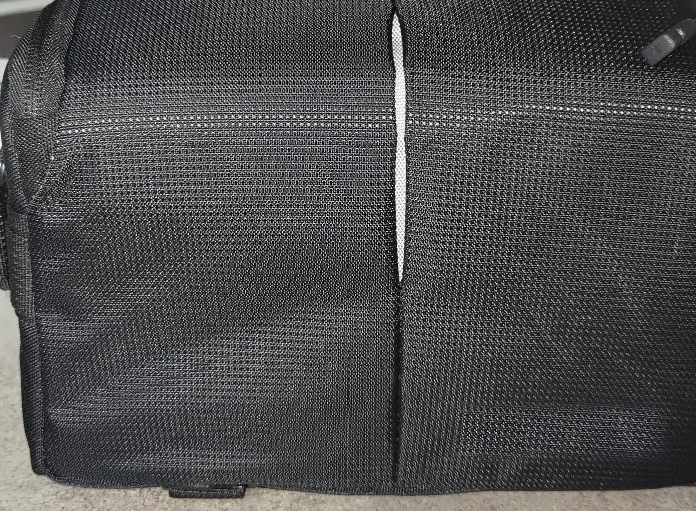 Une sacoche de transport noire, fermée, posée sur un sol clair : toile tissée, une couture verticale au milieu et une sangle réfléchissante le long de l'arête.
