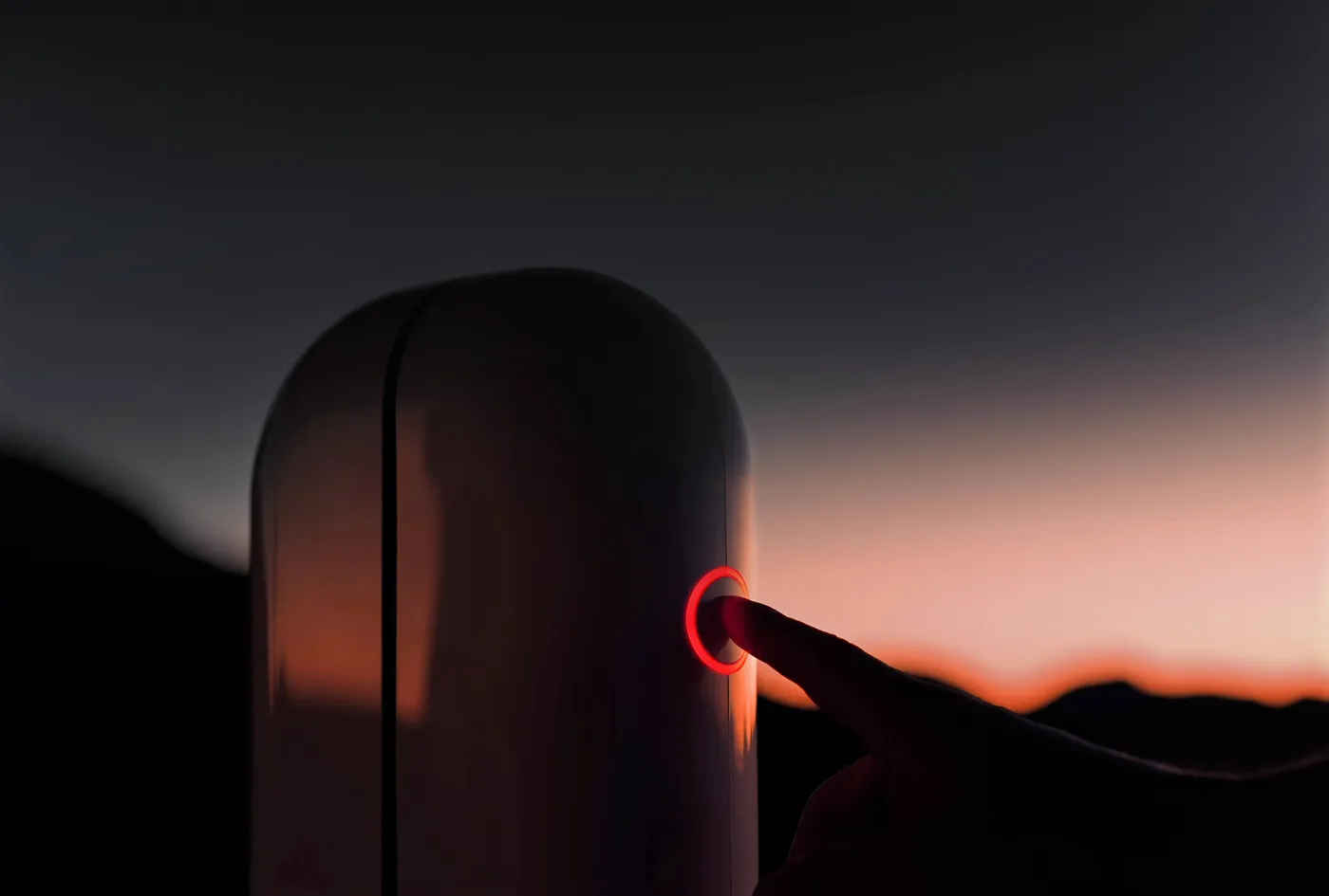 Un doigt presse l'anneau lumineux rouge du Vespera Pro, dehors au crépuscule, devant un horizon orangé.